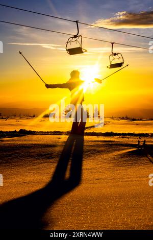 Silhouette of a skier stretching arms against a golden sunset on a snowy mountain slope, with ski lifts in the background. Stock Photo