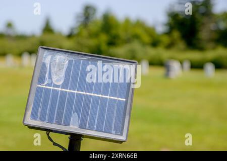 A small damaged solar panel sticks up on a post. Shallow depth of field, trees and a small cemetery behind. Stock Photo