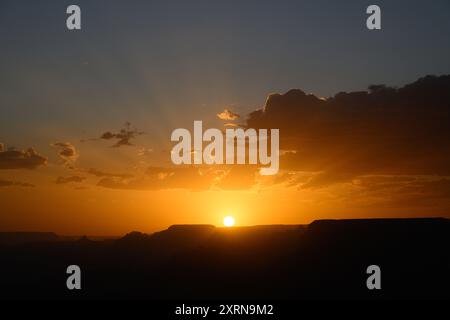 Orange sunrays radiate from setting sun as it touches a ridge beneath clouds in sky Stock Photo