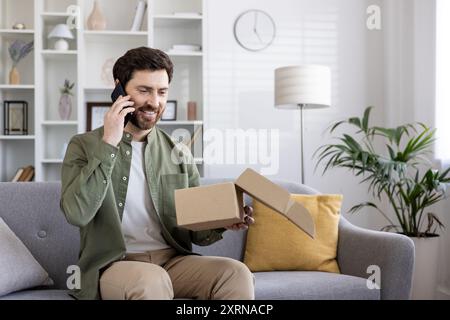 Man smiling and talking on phone while sitting on sofa opening package. Concept of online shopping, home delivery, and communication. Stock Photo