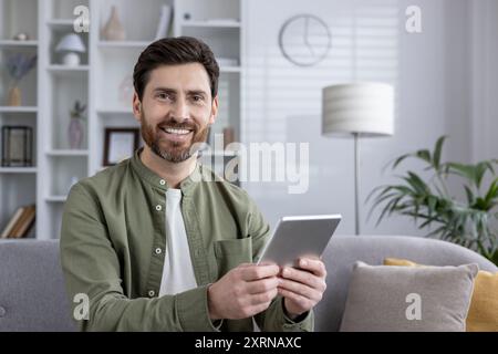 Man sitting on sofa smiling while using tablet at home. Relaxed and cheerful expression, wearing casual clothing, and engaging with device. Bright and cozy living room environment Stock Photo