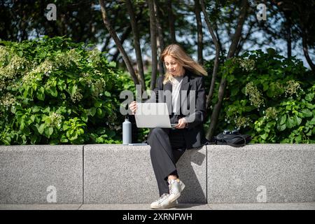 Interested woman freelancer in business suit sits outdoor with laptop on knees distant works  Stock Photo