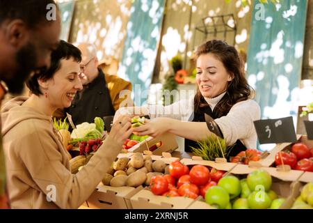 Cheerful diverse couple tasting fresh organic products and shopping at outdoor farmers market stall. Happy female vendor standing behind the farm stand offering customers to taste bio apples. Stock Photo