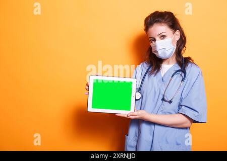 Nurse practitioner with face mask horizontally holding tablet with green screen. Female healthcare specialist grasping digital device displaying isolated copyspace mockup template. Stock Photo