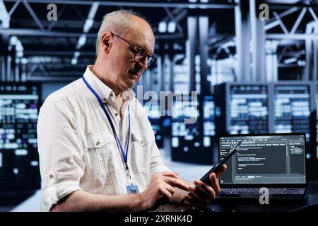 Elderly technician coding in high tech data center using javascript language. Certified specialist updating software on server rigs on laptop terminal, ensuring optimal performance and security Stock Photo