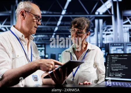 Teamworking colleagues updating server racks, writing intricate binary code scripts on tablet and laptop. Elderly coworkers using programming to fortify cybersecurity in high tech workspace Stock Photo