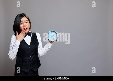 Tired asian woman wearing waitress uniform yawning and covering mouth while holding alarm clock portrait. Sleepy receptionist running late for work, showing time and looking at camera Stock Photo