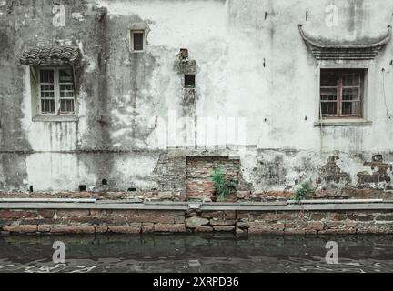 A weathered building facade with windows and a brick wall, reflected in calm water. Stock Photo