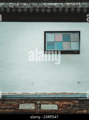 A white plastered wall with a single window in a traditional Chinese building. The window is divided into six panes with different patterned glass. Stock Photo