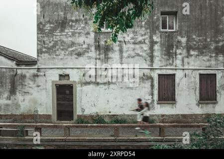 A weathered stone wall in Suzhou, China, with a doorway, windows, and a stone bridge in the foreground. Two figures walk blurry in the distance. Stock Photo