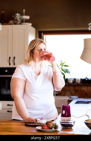 Overweight woman in the kitchen, drinking fresh smoothie. Choosing healthy food, making smoothie at home. Stock Photo