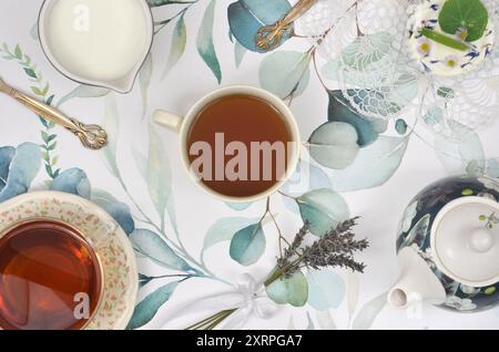 Two cups of tea on a tablecloth with green leaves. Tea pot, milk ina jug, cupcake and two spoons. Top view table. Stock Photo