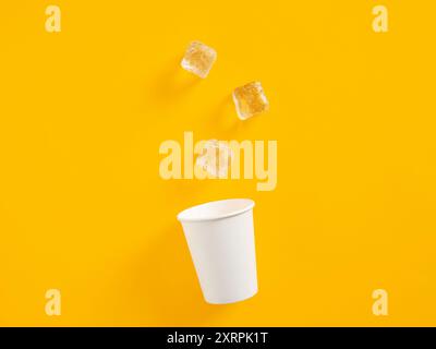 Ice cubes falling into a white paper cup over a yellow background with copy space. Stock Photo