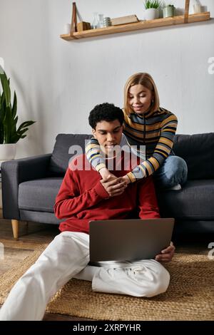 A happy interracial couple relaxes on the floor of their apartment while using a laptop. Stock Photo