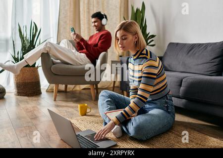 A happy couple relaxes in their modern apartment, one woman on the floor using a laptop while the other man sits in a chair. Stock Photo
