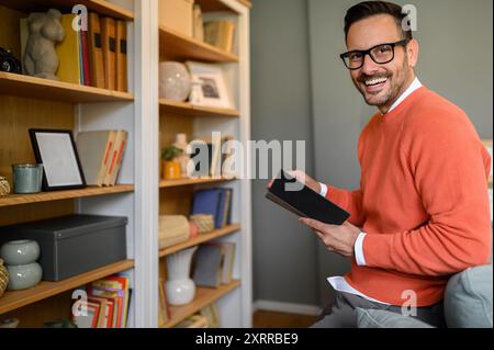 Happy young businessman in orange sweater reading a book while standing by shelf in living room Stock Photo