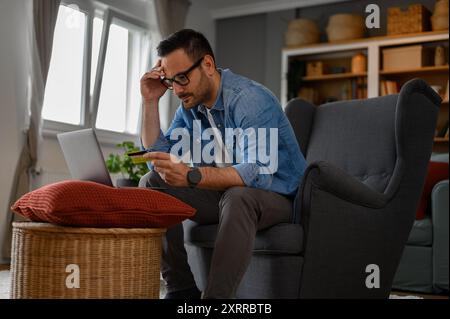 Stressed businessman with credit card paying bills online over laptop while sitting in home office Stock Photo