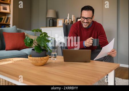 Happy young businessman holding coffee cup and document discussing over video call on laptop at home Stock Photo