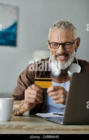 A mature gay man with gray hair and tattoos smiles while holding a credit card and looking at his laptop, working from home. Stock Photo