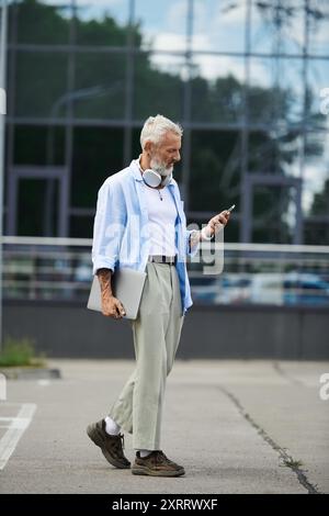 A mature man with a grey beard and tattoos walks down a city street, holding a laptop and checking his phone. Stock Photo