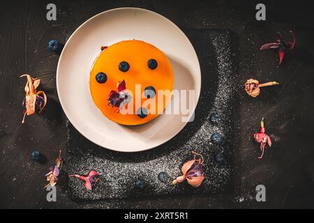 Dessert or cake with layers of chocolate, curd cream, mango jelly, decorated with food dried flowers on a white plate on a black background. Original Stock Photo