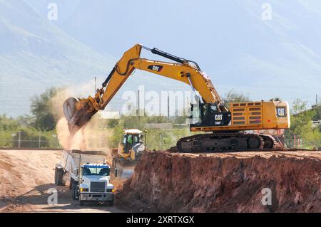 The operator of a yellow large excavator maneuvers its bucket to fill a red and white dump side truck with dirt at a construction site in Pinal County Stock Photo