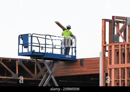 A worker using a scissor lift carrying a beam on to the rooftop of a timber-frame commercial building under construction Stock Photo