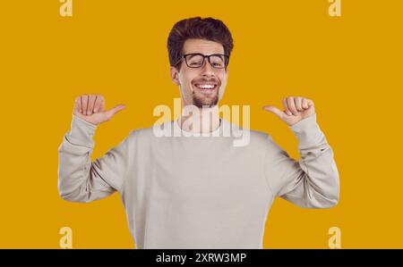 Smiling laughing happy bearded man in glasses pointing thumbs to himself on yellow background. Stock Photo