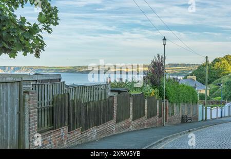 A cobbled road runs downhill and a view to a seaside bay can be seen over a fence. There are chalk cliffs in the distance and a sky withy cloud is abo Stock Photo