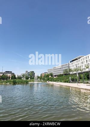 A calm body of water with a clear blue sky above. The water is surrounded by a park and buildings Stock Photo