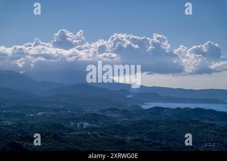 Smoke gently rises from a chimney into the blue sky with rolling white clouds. The coastal green mountains create a serene and picturesque landscape. Stock Photo