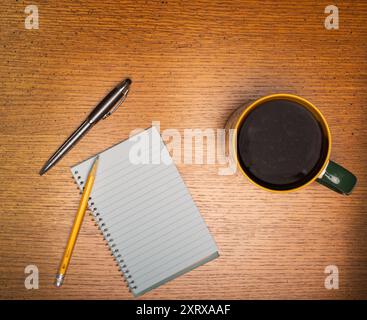 Coffee in gold and green trimmed cup sitting on a rough wood plank table next to plank spiral note pad and pencil Stock Photo