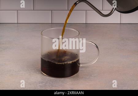 Hot coffee being poured into glass cup on pink tinted counter with white subway tile background. Condensation is forming on the sides of the cup from Stock Photo