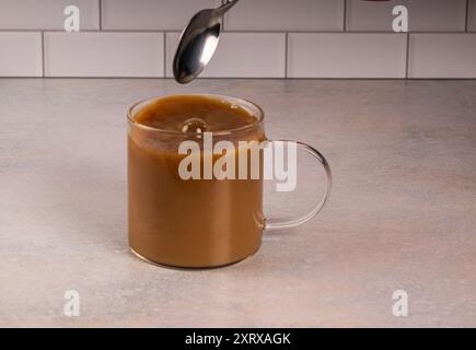 Coffee with creamer in a glass cup swirls and produces a bubble on the surface after being stirred. Cup sits on a pink tinted counter with a white sub Stock Photo