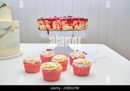 six cupcakes with white cream in pink wrappers, arranged in a row. Behind the cupcakes is a Cheesecake dessert, centered on a white cake stand. Stock Photo