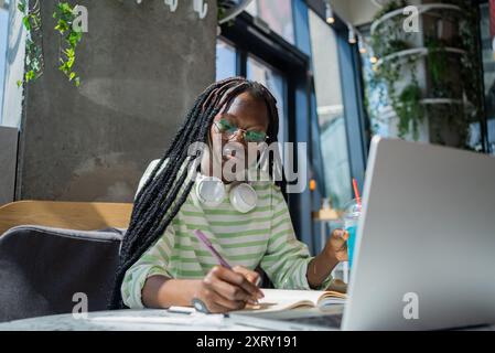 Freelancer african american woman taking notes while working on laptop in trendy cafe and sipping drink, multitasking with headphones and blue drink, remote work and study concept. Focused on tasks. Stock Photo