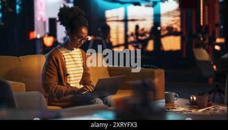 Portrait of Black Creative Young Woman Working on a Laptop in Creative Office at Night. Female Team Lead Smiling While Checking her Team Performance Data, Sending Emails of Feedback Stock Photo