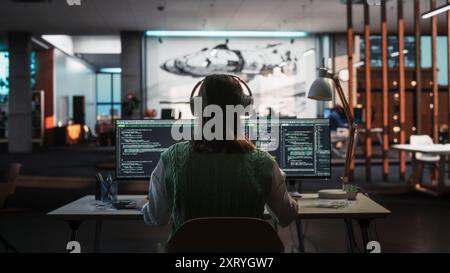 Creative Office: Professional Female Programmer Uses Headphones, Working on Desktop Computer. Focused Coder, Software Engineer Developing App, Video Game. Listening to Podcast or Music. Back View. Stock Photo