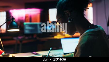 Motivated Black Female Using Laptop and Tablet Computer in Creative Agency in the Evening. Dedicated Woman Working in a Creative Job Position in an Advertising Company Stock Photo