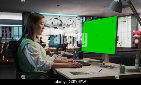 Female Asian Game Programmer Coding On Desktop Computer With Green Screen Chromakey On Display In Game Design Studio Office. Stylish Woman Does Gameplay Engineering For New Immersive 3D RPG Video Game Stock Photo