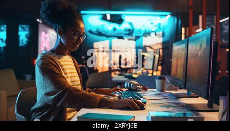 Skillful Female Designer Adding 3D Model Elements while Working on a New Video Game on Desktop Computer in Creative Office During the Evening. Girl Satisfied with her Work. Graphic Designer Concept Stock Photo