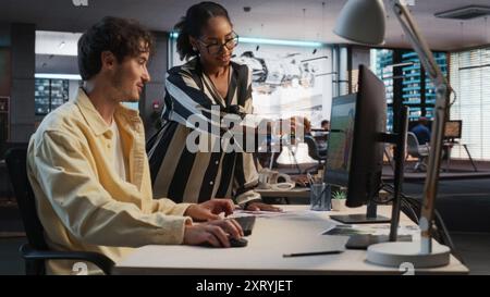 Caucasian Male Game Designer Using Desktop Computer With 3D Modelling Software And Talking to Black Female Project Manager in Game Development Studio Office. Diverse Colleagues Discussing New RPG Game Stock Photo