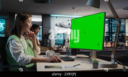 Female Asian Game Programmer Coding On Desktop Computer With Green Screen Chromakey On Display In Game Design Studio Office. Stylish Woman Does Gameplay Engineering For New Immersive 3D RPG Video Game Stock Photo