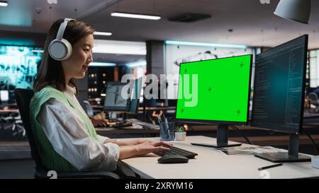 Female Asian Game Programmer Coding On Desktop Computer With Green Screen Chromakey On Display In Game Design Studio Office. Focused Woman Does Gameplay Engineering For New Immersive 3D RPG Video Game Stock Photo