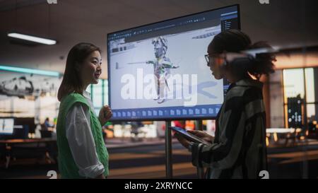 Black Female Game Designer Talking to Asian Female Project Manager And Standing Next To TV Set With 3D Game Character On Screen. Diverse Colleagues Discussing New RPG Game in Office Stock Photo