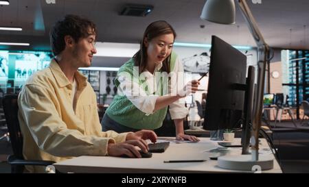 Caucasian Male Game Designer Using Desktop Computer With 3D Modelling Software And Talking to Asian Female Project Manager in Game Development Studio Office. Diverse Colleagues Discussing New RPG Game Stock Photo