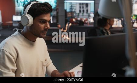 Male Indian Game Programmer Coding On Desktop Computer In Game Design Studio Diverse Office. Focused Man Writes Lines Of Code, Does Gameplay Engineering For New Immersive 3D Survival Video Game Stock Photo