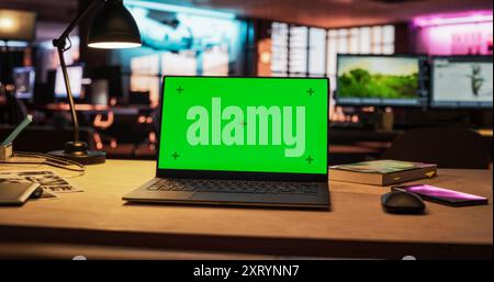 Mock-up Green Screen on a Laptop Standing on the top of a Wooden Table. In the Background Stylish Modern Office Studio in the Evening with Neon Lights. Desk of Creative Designer Stock Photo