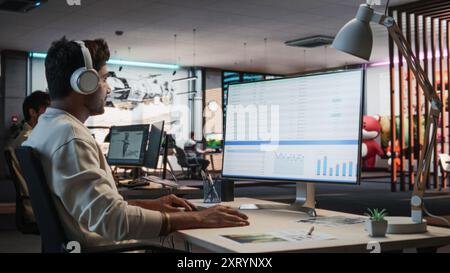 Indian Male Manager Using Desktop Computer With Analytics Software wth Financial Reports. Man Working In Game Development Company Diverse Office Stock Photo