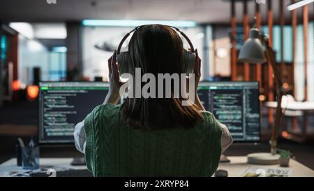 Creative Office: Professional Female Programmer Uses Headphones, Working on Desktop Computer. Focused Developer, Software Engineer Developing App, Video Game. Listening to Podcast or Music. Back View. Stock Photo